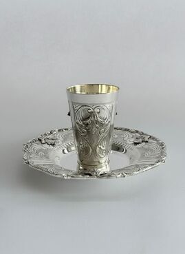Silver cup with intricate designs on a matching saucer against a light gray background