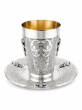 An ornate sterling silver Kiddush cup with a gold interior, featuring intricate floral engravings and side handles, shown on a matching engraved silver saucer by hadad bros silverware