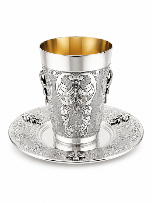 An ornate sterling silver Kiddush cup with a gold interior, featuring intricate floral engravings and side handles, shown on a matching engraved silver saucer by hadad bros silverware