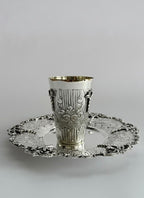 Silver kiddush cup with intricate designs on a matching saucer against a white background
