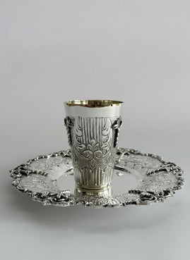 Silver kiddush cup with intricate designs on a matching saucer against a white background